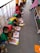 A group of children is sitting on the floor in a line, each engaged in a coloring activity. They are using colorful crayons and focusing on filling in pictures. The scene appears to be in an outdoor or semi-outdoor walkway area, with a railing on one side. The children are wearing brightly colored clothing, which might be uniforms.