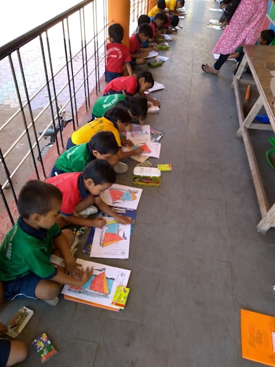 A group of children is sitting on the floor in a line, each engaged in a coloring activity. They are using colorful crayons and focusing on filling in pictures. The scene appears to be in an outdoor or semi-outdoor walkway area, with a railing on one side. The children are wearing brightly colored clothing, which might be uniforms.