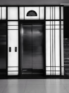A modern elevator is centered between illuminated panels with a minimalist, geometric design. The elevator doors are metallic and closed, with control buttons on either side. Above the doors, a display shows the number 3.
