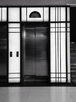 A modern elevator is centered between illuminated panels with a minimalist, geometric design. The elevator doors are metallic and closed, with control buttons on either side. Above the doors, a display shows the number 3.