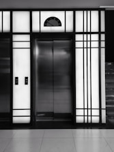 A modern elevator is centered between illuminated panels with a minimalist, geometric design. The elevator doors are metallic and closed, with control buttons on either side. Above the doors, a display shows the number 3.