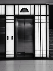 A modern elevator is centered between illuminated panels with a minimalist, geometric design. The elevator doors are metallic and closed, with control buttons on either side. Above the doors, a display shows the number 3.