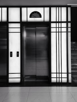 A modern elevator is centered between illuminated panels with a minimalist, geometric design. The elevator doors are metallic and closed, with control buttons on either side. Above the doors, a display shows the number 3.