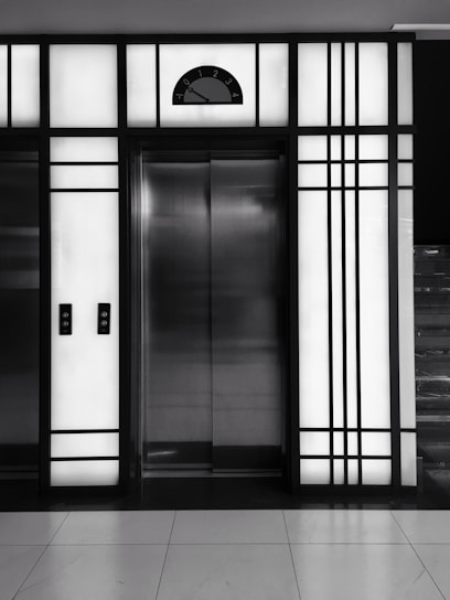 A modern elevator is centered between illuminated panels with a minimalist, geometric design. The elevator doors are metallic and closed, with control buttons on either side. Above the doors, a display shows the number 3.