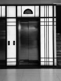 A modern elevator is centered between illuminated panels with a minimalist, geometric design. The elevator doors are metallic and closed, with control buttons on either side. Above the doors, a display shows the number 3.