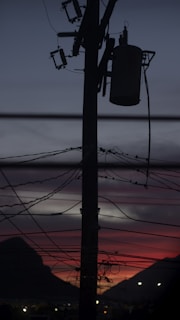 A silhouette of an electricity pole with multiple wires and transformers against a backdrop of a dramatic sunset with red and purple hues in the sky, flanked by dark mountains.