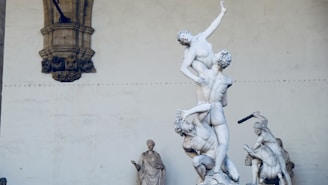 A classical sculpture depicting several figures in a dramatic pose, with one figure being lifted by another. The setting appears to be an outdoor historical space with an arched wall as a backdrop. Additional sculptures are displayed in the background.