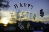 Colorful letters spell out 'Happy Easter' against a window with a view of a sunset or sunrise in the background. Decorative Easter eggs surround the text, and there is a blurred silhouette of trees and buildings.