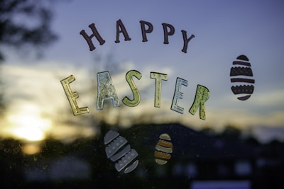 Colorful letters spell out 'Happy Easter' against a window with a view of a sunset or sunrise in the background. Decorative Easter eggs surround the text, and there is a blurred silhouette of trees and buildings.