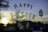 Colorful letters spell out 'Happy Easter' against a window with a view of a sunset or sunrise in the background. Decorative Easter eggs surround the text, and there is a blurred silhouette of trees and buildings.