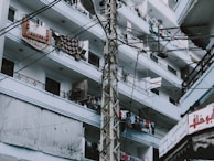 An urban residential building with multiple floors and balconies is shown. The balconies are filled with various items such as hanging clothes and rugs. A utility pole with numerous cables runs across the image, and there's a sign with Arabic script in the lower right corner.