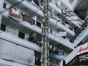 An urban residential building with multiple floors and balconies is shown. The balconies are filled with various items such as hanging clothes and rugs. A utility pole with numerous cables runs across the image, and there's a sign with Arabic script in the lower right corner.