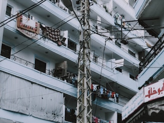 An urban residential building with multiple floors and balconies is shown. The balconies are filled with various items such as hanging clothes and rugs. A utility pole with numerous cables runs across the image, and there's a sign with Arabic script in the lower right corner.