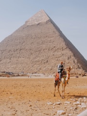 A majestic pyramid rises prominently in the background with a man riding a camel in the foreground. The scene is set in a desert environment, and there are several people visible in the distance, adding scale to the vastness of the pyramid. The sunlight casts subtle shadows, enhancing the texture of the pyramid's stones.
