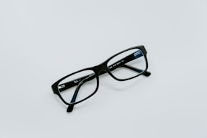 A pair of black rectangular eyeglasses lies on a plain light gray surface. The frame is sleek and modern, with clear lenses reflecting a hint of blue light. The glasses are positioned at an angle, casting a subtle shadow.