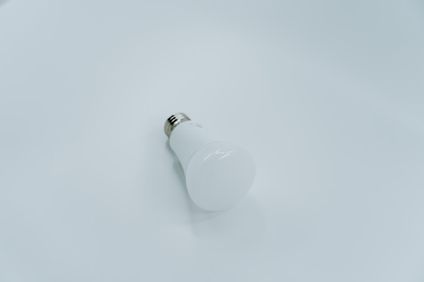 A white LED light bulb lies on a smooth, light gray surface. The bulb is positioned at an angle with the metallic base visible, creating a minimalist and clean look.