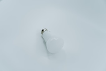 A white LED light bulb lies on a smooth, light gray surface. The bulb is positioned at an angle with the metallic base visible, creating a minimalist and clean look.