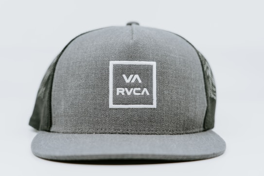 A gray baseball cap featuring a structured front panel with a mesh back. The front is adorned with a square logo containing the letters VA and RVCA in white stitching.