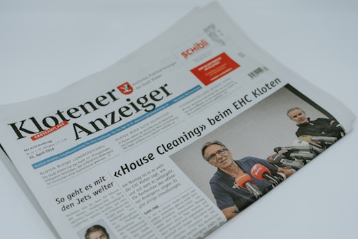 A folded newspaper with the header 'Klotener Anzeiger' is prominently displayed. Below the header, there is an article headline in bold about 'House Cleaning' at EHC Kloten. A photo beneath the headline shows a person wearing glasses speaking at a press conference, surrounded by microphones from various news organizations.