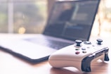 A close-up of a white gaming controller is placed on a wooden surface alongside a laptop that is slightly blurred in the background, showing a screen with text or code.