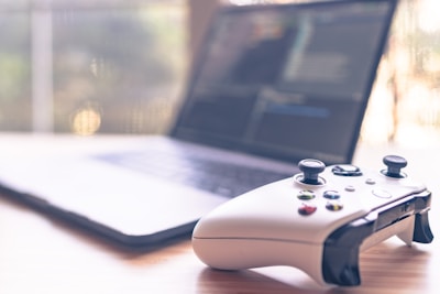 A close-up of a white gaming controller is placed on a wooden surface alongside a laptop that is slightly blurred in the background, showing a screen with text or code.