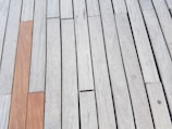A pattern of wooden planks laid parallel to each other, predominantly in a light gray tone with one plank in a contrasting reddish-brown hue.