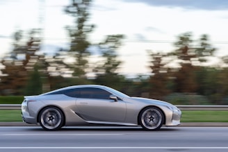 A sleek silver sports car is moving swiftly along a road, with blurred trees and landscape in the background indicating speed. The car's modern design and shiny exterior suggest a high-performance vehicle, exuding a sense of luxury and precision.