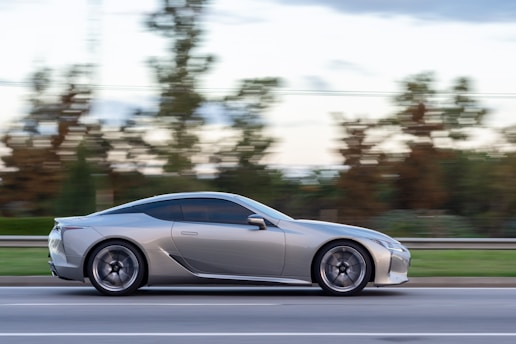 A sleek silver sports car is moving swiftly along a road, with blurred trees and landscape in the background indicating speed. The car's modern design and shiny exterior suggest a high-performance vehicle, exuding a sense of luxury and precision.