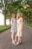Two women are standing on a tree-lined path. The woman on the left is wearing a light blue floral dress and white high-heeled sandals. The woman on the right is wearing a sleeveless, beige dress with vertical stripes and tan high-heeled sandals. They are smiling and positioned closely together, with one woman resting her hand on the other’s shoulder. The background features tall trees with green foliage, a grassy area, and a wooden fence along the left side.