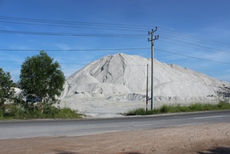 A large pile of white sand or limestone is situated in an open area, with a road and some greenery in the foreground. Power lines run across the scene, supported by utility poles. The sky is clear and blue, contributing to a serene atmosphere.