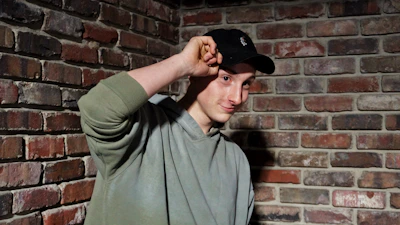 Close-up of hands adjusting a snapback cap with the riotvibe logo, set against a gritty urban wall.