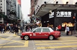 A red taxi is seen driving across a busy city street with numerous pedestrians crossing at the intersection. The area is densely populated with high-rise buildings and stores, including a visible Lukfook Jewellery shop. The atmosphere is bustling, with people engaged in various activities.