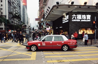A red taxi is seen driving across a busy city street with numerous pedestrians crossing at the intersection. The area is densely populated with high-rise buildings and stores, including a visible Lukfook Jewellery shop. The atmosphere is bustling, with people engaged in various activities.