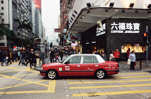 A red taxi is seen driving across a busy city street with numerous pedestrians crossing at the intersection. The area is densely populated with high-rise buildings and stores, including a visible Lukfook Jewellery shop. The atmosphere is bustling, with people engaged in various activities.