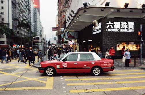 A red taxi is seen driving across a busy city street with numerous pedestrians crossing at the intersection. The area is densely populated with high-rise buildings and stores, including a visible Lukfook Jewellery shop. The atmosphere is bustling, with people engaged in various activities.