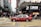 A red taxi is seen driving across a busy city street with numerous pedestrians crossing at the intersection. The area is densely populated with high-rise buildings and stores, including a visible Lukfook Jewellery shop. The atmosphere is bustling, with people engaged in various activities.