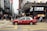 A red taxi is seen driving across a busy city street with numerous pedestrians crossing at the intersection. The area is densely populated with high-rise buildings and stores, including a visible Lukfook Jewellery shop. The atmosphere is bustling, with people engaged in various activities.