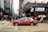 A red taxi is seen driving across a busy city street with numerous pedestrians crossing at the intersection. The area is densely populated with high-rise buildings and stores, including a visible Lukfook Jewellery shop. The atmosphere is bustling, with people engaged in various activities.