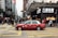 A red taxi is seen driving across a busy city street with numerous pedestrians crossing at the intersection. The area is densely populated with high-rise buildings and stores, including a visible Lukfook Jewellery shop. The atmosphere is bustling, with people engaged in various activities.