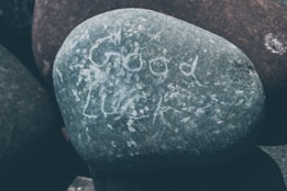 A smooth, round stone with the words 'Good Luck' etched onto its surface. It is surrounded by other similar stones, showcasing a natural and earthy texture.