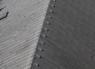 A close-up view of a wooden roof with a pattern of overlapping shingles. The texture and grain of the wood are visible, and there is a triangular vent or window set into the roof.