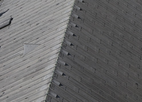 A close-up view of a wooden roof with a pattern of overlapping shingles. The texture and grain of the wood are visible, and there is a triangular vent or window set into the roof.