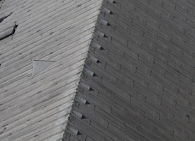 A close-up view of a wooden roof with a pattern of overlapping shingles. The texture and grain of the wood are visible, and there is a triangular vent or window set into the roof.