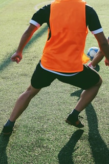 A person wearing an orange sports vest and black shorts is playing soccer on a grassy field. The person is in motion with a slight crouch, and there is a soccer ball in front of them.