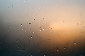 Droplets of water are scattered across a transparent surface, such as glass, with a softly blurred background featuring warm, muted tones.