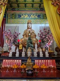 An ornate altar featuring a large golden Buddha statue surrounded by smaller statues. The altar is adorned with vibrant pink flowers, offerings of oranges, and red candles. Intricately decorated walls and ceiling frame the scene, featuring traditional patterns and bright colors. Two red lanterns are lit, adding to the serene and sacred atmosphere.