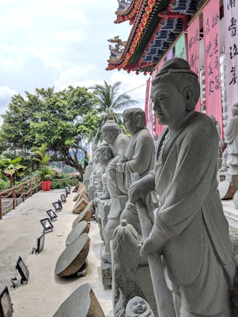 A row of stone statues depicting serene figures stands beside a temple adorned with colorful banners featuring intricate characters. The ornate, traditional Chinese architectural elements of the temple are prominently displayed. Lush greenery, including trees and a palm, borders the pathway which is lined with artificial lighting.