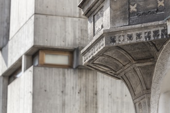 Detailed architectural features of a concrete structure including geometric patterns and reliefs. The forefront showcases a prominent overhang with intricate designs and textures, set against a background of additional concrete surfaces with a small, recessed window.
