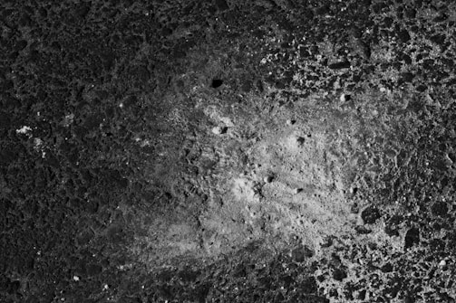 A textured surface with a rough, uneven pattern, resembling a lunar landscape or a close-up view of volcanic rock. The surface features various shades of gray, with small craters and an irregular distribution of lighter and darker areas.