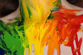 Brightly colored paint is smeared across a person's back and shoulders blending green, yellow, orange, and red. The paint appears thick and textured, creating an abstract and vibrant visual effect.
